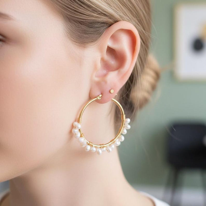 May include: Gold hoop earrings with small, white pearls dangling from the bottom. The earrings are worn on a person's ear, with a small gold stud earring visible above the hoop. The hoops are a classic, elegant jewelry piece.