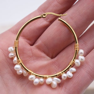 May include: Gold-tone hoop earrings adorned with small, white, teardrop-shaped pearls. The pearls are clustered and suspended from the bottom of the hoops. The hoops are secured with a gold-colored clasp.