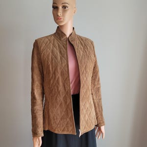 May include: A tan quilted jacket with a stand-up collar and a zipper closure. The jacket is displayed on a mannequin, paired with a pink top and a black skirt. The jacket's diamond pattern adds texture to the garment.