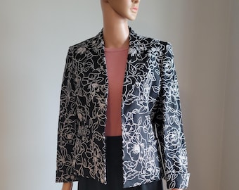 May include: A black blazer with a white floral pattern is displayed on a mannequin. The blazer has long sleeves and a classic cut. The mannequin is also wearing a pink top and a black skirt, creating a coordinated outfit.