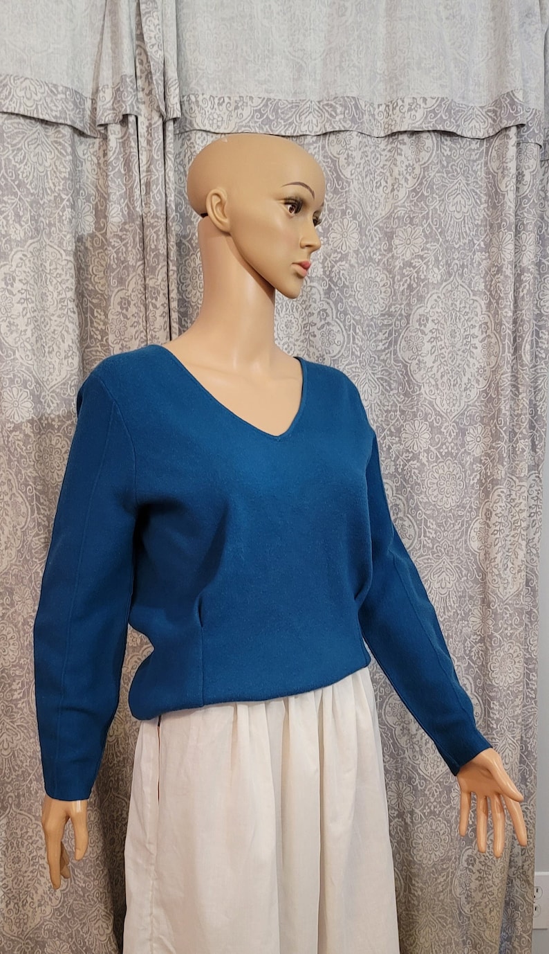 Pu&ograve; includere: Un maglione blu teal a collo a V con maniche lunghe. Il maglione &egrave; su un manichino che indossa una gonna bianca.