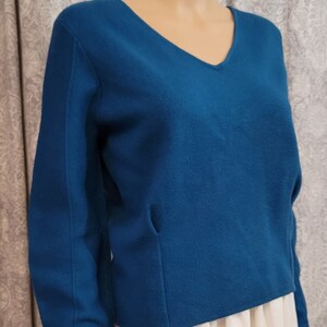 Pu&ograve; includere: Un maglione blu teal a maniche lunghe con scollo a V. Il maglione &egrave; realizzato in un tessuto morbido e lavorato a maglia.