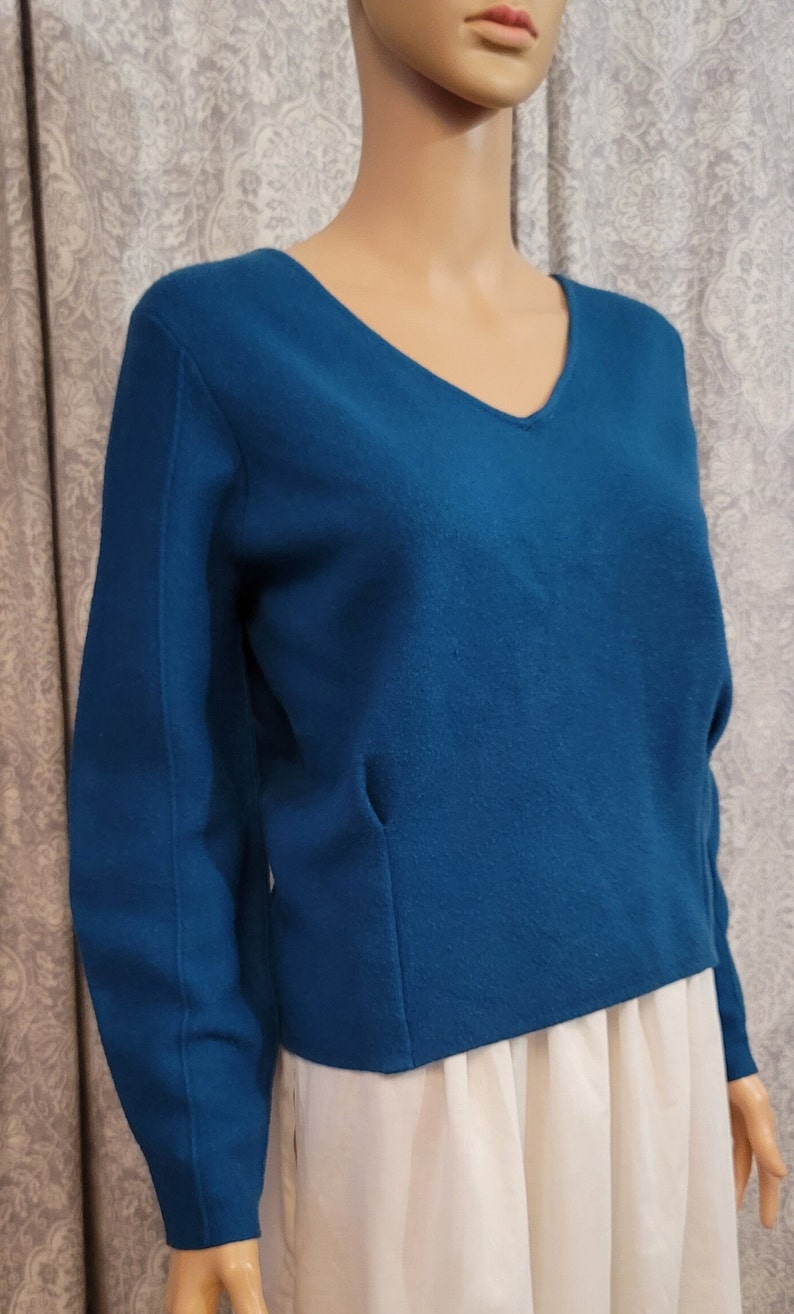 Pu&ograve; includere: Un maglione blu teal a maniche lunghe con scollo a V. Il maglione &egrave; realizzato in un tessuto morbido e lavorato a maglia.