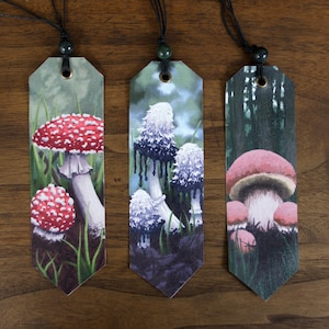 May include: Three decorative bookmarks featuring mushroom illustrations. The first bookmark displays a red and white spotted mushroom. The second bookmark shows white and black mushrooms. The third bookmark has a pink mushroom. Each bookmark has a black cord.