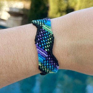 May include: A colorful friendship bracelet with a woven pattern. The bracelet features a combination of blue, green, purple, and black threads. The bracelet is worn on a wrist, with a blurred background of trees and a pool.