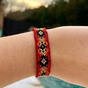 May include: A woven friendship bracelet with a geometric pattern. The bracelet features a red base with orange, black, and white diamond-shaped designs. The bracelet is worn on a wrist, showcasing the vibrant colors and intricate weaving.