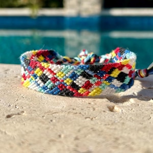 May include: A colorful friendship bracelet with a woven pattern. The bracelet features a mix of red, yellow, black, white, and blue threads. The bracelet is approximately 0.75 inches wide and has a tassel at the end.