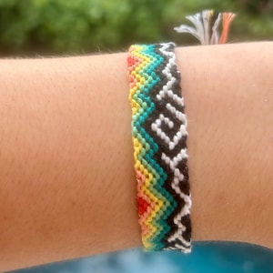 May include: A colorful friendship bracelet with a chevron pattern. The bracelet features a gradient of colors, including red, yellow, green, and black and white. The bracelet is handmade and has a woven design.