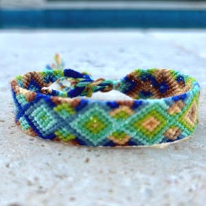 May include: A handmade friendship bracelet with a woven pattern. The bracelet features a repeating diamond design in shades of blue, green, brown, and tan. The bracelet is displayed on a textured, light-colored surface.
