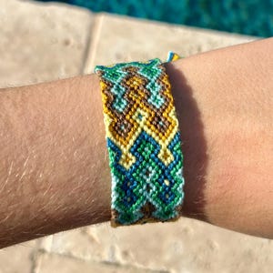 May include: A handmade friendship bracelet with a woven pattern. The bracelet features a combination of green, blue, yellow, and brown threads. The design is intricate and geometric, with a textured appearance. The bracelet is worn on a wrist.