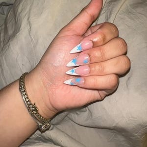 May include: Close-up of a hand with long, white, stiletto-shaped nails with a blue star design on each nail.