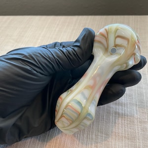 May include: A hand-blown glass pipe with a white base and colorful swirls of peach, green, and yellow. The pipe has a rounded bowl and a long, slender stem. The pipe is held by a gloved hand.