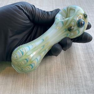 May include: A hand-blown glass pipe with a swirling pattern of light green, blue, and yellow. The pipe has a rounded bowl and a long, curved stem. Three small, decorative glass beads are on the bowl.
