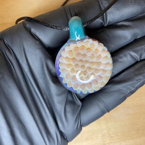 May include: A handcrafted glass pendant with a black cord. The pendant features a teal top, a light yellow and white patterned center, and a purple base. The design includes a white, curved accent.
