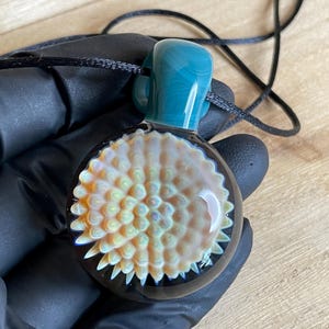 May include: A handcrafted glass pendant with a teal bail and a round, textured body. The body features a detailed, layered design in shades of white, yellow, and orange, set against a dark background. The pendant hangs from a black cord.