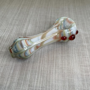 Pode incluir: Um cachimbo de vidro soprado à mão com um padrão de redemoinho de branco, laranja, verde e azul. O cachimbo apresenta três detalhes em vidro vermelho na extremidade da tigela. O cachimbo foi projetado para fumar.