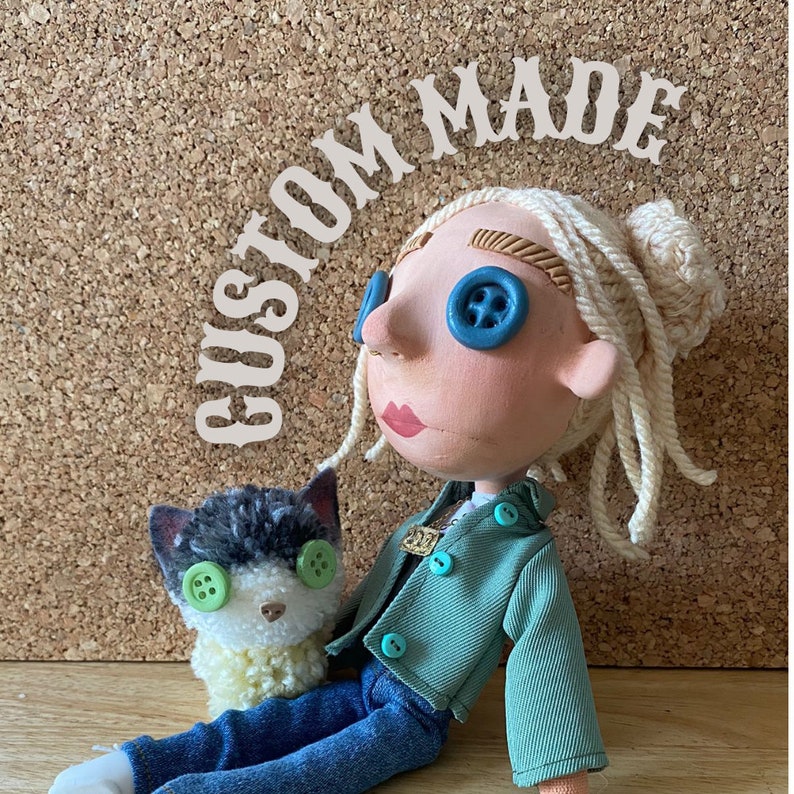 May include: A handmade doll with button eyes and a matching cat. The doll has blonde yarn hair and is wearing a green jacket and blue jeans. The cat has green button eyes and a fluffy yellow and white collar. The words "CUSTOM MADE" are arched above the doll.