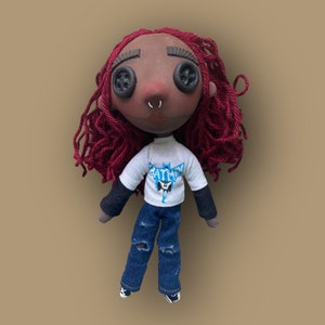 May include: Handmade doll with button eyes, red yarn hair, and a nose ring. The doll wears a white t-shirt with a Batman logo, a black long-sleeve shirt, and distressed blue jeans. The doll's face is painted with a dark skin tone.
