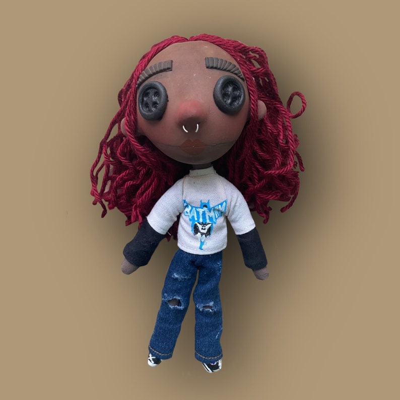 May include: Handmade doll with button eyes, red yarn hair, and a nose ring. The doll wears a white t-shirt with a Batman logo, a black long-sleeve shirt, and distressed blue jeans. The doll's face is painted with a dark skin tone.