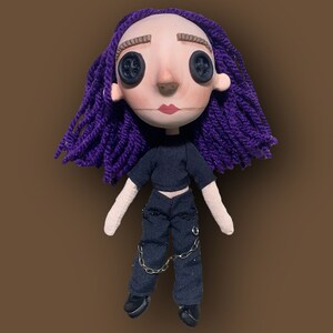 May include: A handmade doll with a pale face, button eyes, and purple yarn hair. The doll wears a black crop top, black pants with chain accents, and black shoes. The doll has a neutral expression.