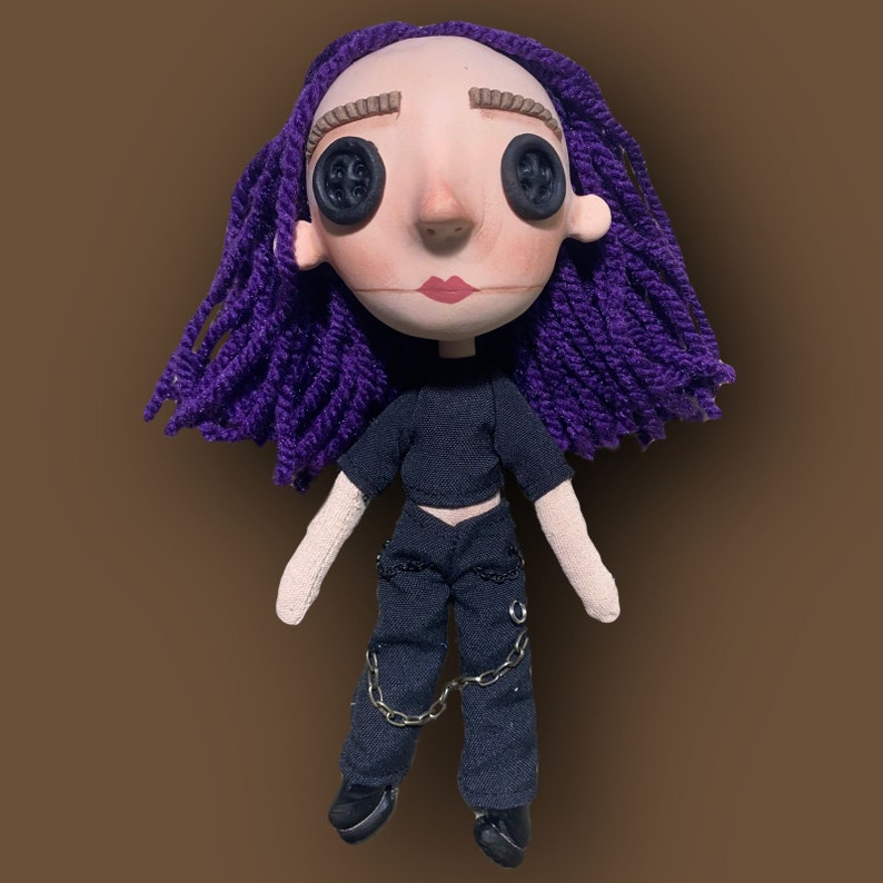 May include: A handmade doll with a pale face, button eyes, and purple yarn hair. The doll wears a black crop top, black pants with chain accents, and black shoes. The doll has a neutral expression.