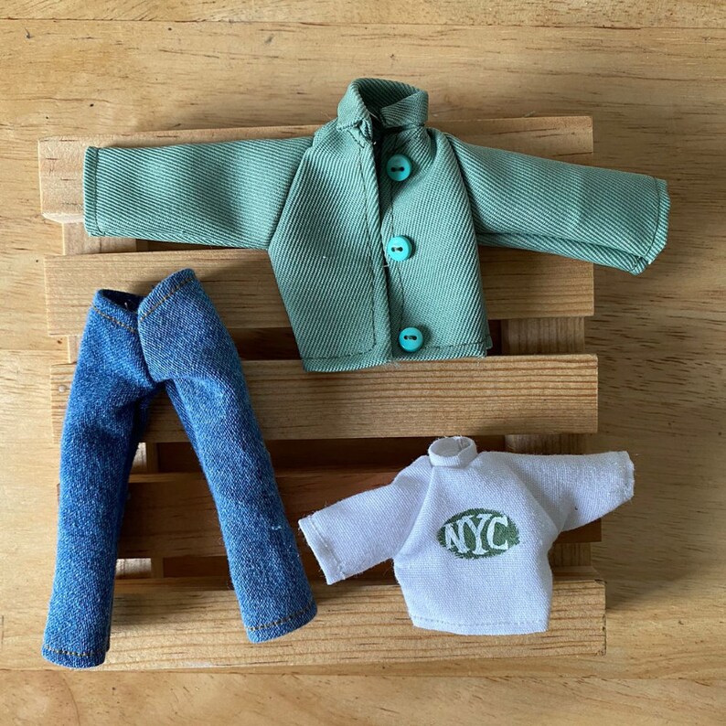 May include: A miniature outfit featuring a green jacket with turquoise buttons, blue denim jeans, and a white sweatshirt with "NYC" printed in green. The clothing is arranged on a wooden slatted surface.