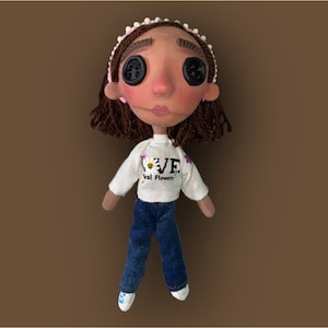 May include: A handmade doll with button eyes, wearing a white sweatshirt with "LOVE real flowers" text, blue jeans, and white shoes. The doll has brown hair, a pink headband, and rosy cheeks. The doll is a unique, handcrafted item.