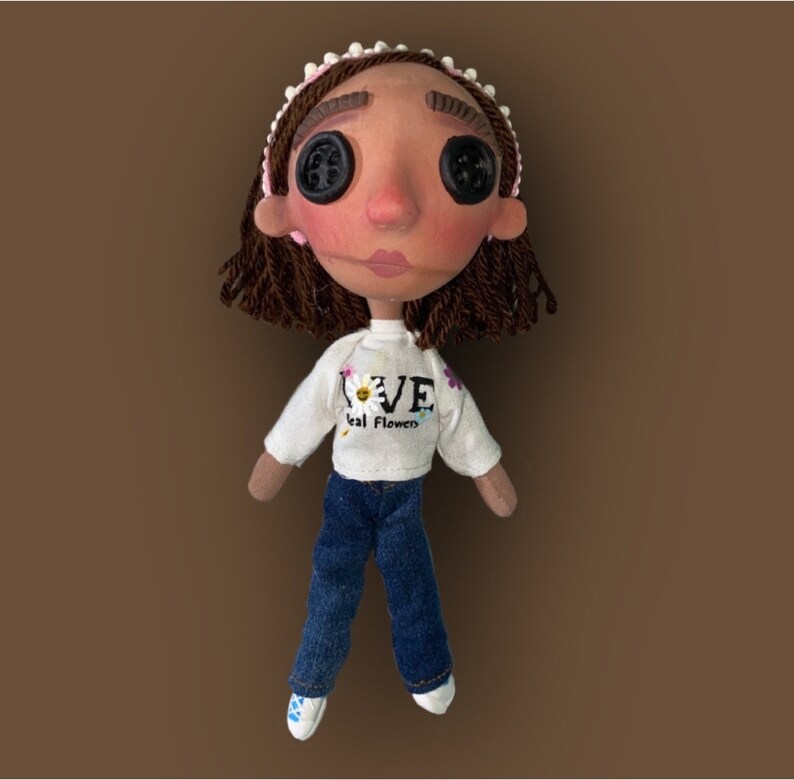 May include: A handmade doll with button eyes, wearing a white sweatshirt with "LOVE real flowers" text, blue jeans, and white shoes. The doll has brown hair, a pink headband, and rosy cheeks. The doll is a unique, handcrafted item.