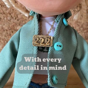 May include: A doll wearing a light green jacket with turquoise buttons, a white t-shirt, and blue jeans. A gold-colored necklace with the number "222" is visible. The image includes the text "With every detail in mind."