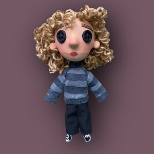 May include: A handmade doll with button eyes, curly blonde hair, and red lips. The doll wears a gray and blue striped sweater, black pants, and sneakers. The doll is set against a muted purple background.