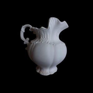 Op de afbeelding: Een witte keramische kan met een gerimpelde rand en een sierlijk handvat. De kan heeft een bolvormig lichaam en een decoratieve basis. De achtergrond is effen zwart, wat de vorm en details van de kan benadrukt.