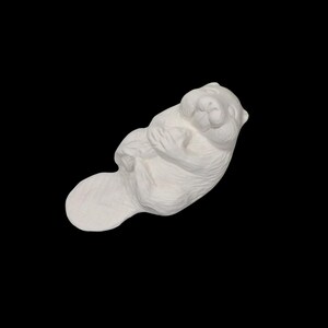 Puede incluir: Escultura de cerámica blanca de un castor en una pose relajada. El castor está tumbado sobre su espalda con los brazos cruzados sobre el pecho. La escultura tiene un acabado liso y mate sobre un fondo negro.