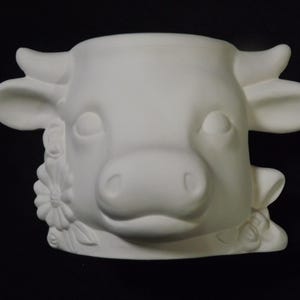 May include: A white ceramic planter shaped like a cow's head. The planter features detailed facial features, including eyes, a nose, and ears. Floral accents are sculpted on the sides. The cow planter is designed to hold plants or flowers.