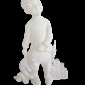 May include: A white ceramic figurine of a young child standing next to a stack of four alphabet blocks. The child wears a long-sleeved shirt and pants with a belt, and holds a small toy. The blocks feature the letters W, A, S, and M in relief.