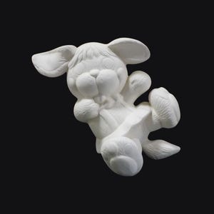 May include: A white ceramic figurine of a cartoon dog in a playful pose. The dog has large ears, a round face, and is wearing a small bow. The figurine is unpainted and ready for customization. The background is black.