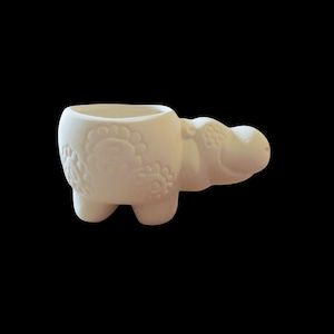 Puede incluir: Una maceta de cerámica con forma de hipopótamo de color crema, con detalles de patrones florales en el lateral. La maceta tiene una pequeña abertura para plantas y mide aproximadamente 15 cm de largo.