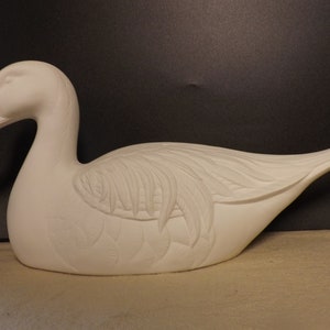 May include: A white ceramic duck figurine with detailed feather carvings on its wings and body. The duck is in a resting position, with its head turned slightly. The figurine is a decorative item.