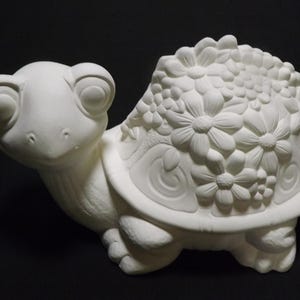 Può includere: Statuetta di tartaruga in ceramica non verniciata, con un elaborato motivo floreale sul guscio. La tartaruga ha grandi occhi espressivi ed è posizionata su una superficie scura. Pronta per essere dipinta.