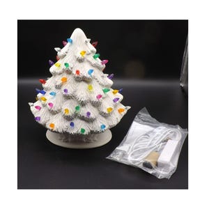 May include: A white ceramic Christmas tree with colorful lights. The tree has a textured surface and is on a round base. A plastic bag containing a power cord and other components is next to the tree.