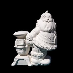 May include: A white ceramic Santa Claus figurine holding a small stove. The Santa figure has a smiling face, a long beard, and a fur-trimmed coat. The stove has a rectangular shape with vertical lines.