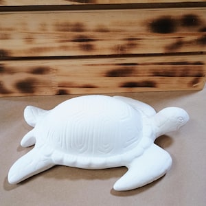 Puede incluir: Una figura de tortuga marina de cerámica blanca que presenta un caparazón detallado con patrones geométricos en relieve, cuatro aletas y una cabeza extendida hacia adelante. La superficie es lisa y mate, diseñada para pintar o decorar.