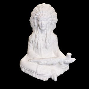 Pode incluir: Uma escultura de cerâmica branca de um chefe nativo americano sentado. A figura usa um cocar de penas e segura um cachimbo. A escultura tem traços faciais detalhados e está sobre um fundo preto.