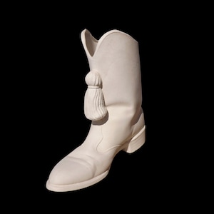 May include: A white ceramic cowboy boot with a decorative tassel. The boot has a pointed toe, a mid-height heel, and a curved top. The background is solid black.