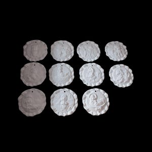 Puede incluir: Una colección de doce adornos blancos, circulares, con bordes festoneados. Cada adorno presenta un diseño en bajorrelieve que representa varias figuras. Los adornos están dispuestos sobre un fondo negro.