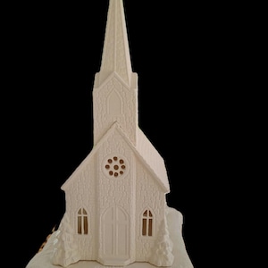 Può includere: Una statuetta di chiesa in ceramica bianca con un alto campanile, un rosone centrale, finestre ad arco e un ingresso a doppia porta. La struttura poggia su una base testurizzata con alberi sempreverdi scolpiti ai lati della scalinata.