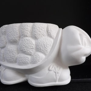 Puede incluir: Una maceta de cerámica blanca con forma de tortuga, con un caparazón texturizado y una cara sonriente. La tortuga tiene una amplia abertura para plantar y es adecuada para plantas pequeñas o suculentas.