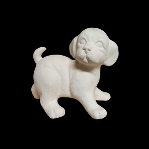 Könnte beinhalten: Eine weiße Keramik-Welpenfigur in spielerischer Pose. Der Welpe hat große, schlaffe Ohren und einen kleinen Schwanz. Die Figur ist unbemalt, bereit zur individuellen Gestaltung und vor einem schwarzen Hintergrund platziert.