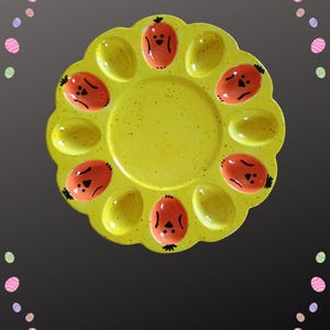 May include: A yellow ceramic Easter egg plate with a scalloped edge. The plate has egg-shaped indentations and is decorated with orange egg-shaped faces. The plate is a bright, cheerful addition to any Easter celebration.