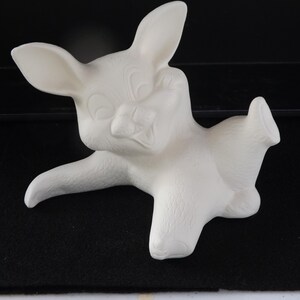 May include: A white ceramic figurine of a cartoon deer in a playful pose. The deer has large ears, a smiling face, and is lying down with one leg extended. The figurine is unpainted and has a smooth surface.
