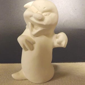 May include: A ceramic figurine of a cartoon ghost, painted in a matte white finish. The ghost has a smiling expression, with arms outstretched and a small tail. The figurine is standing on a neutral-colored surface.